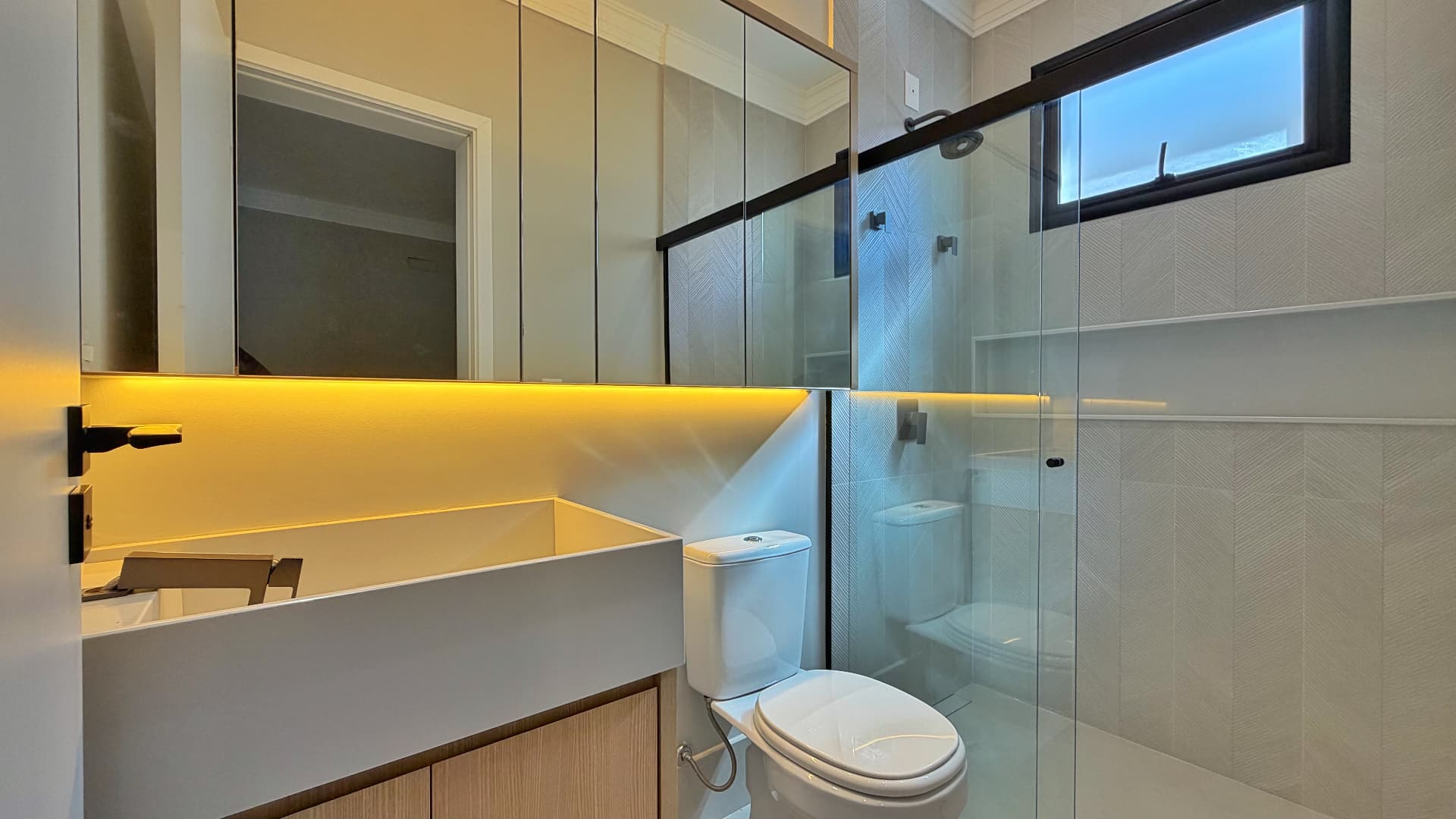 Banheiro da suíte com bancada iluminada, box de vidro e janela basculante