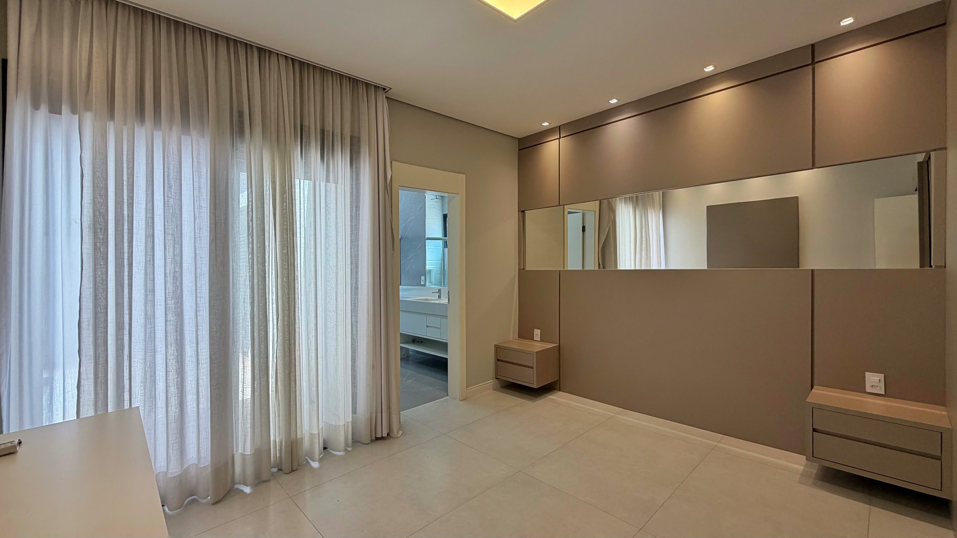 Suíte iluminada com cortinas, painel amadeirado e banheiro integrado